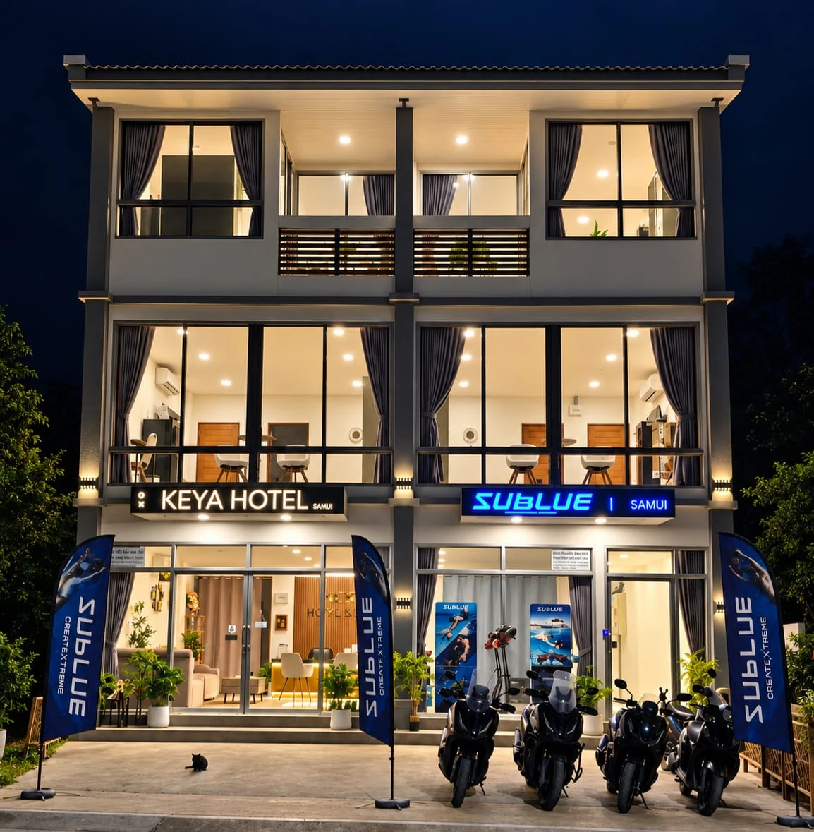 The Keya Hotel Samui building lit up at nightfall, with the Sublue shop and the rental scooters