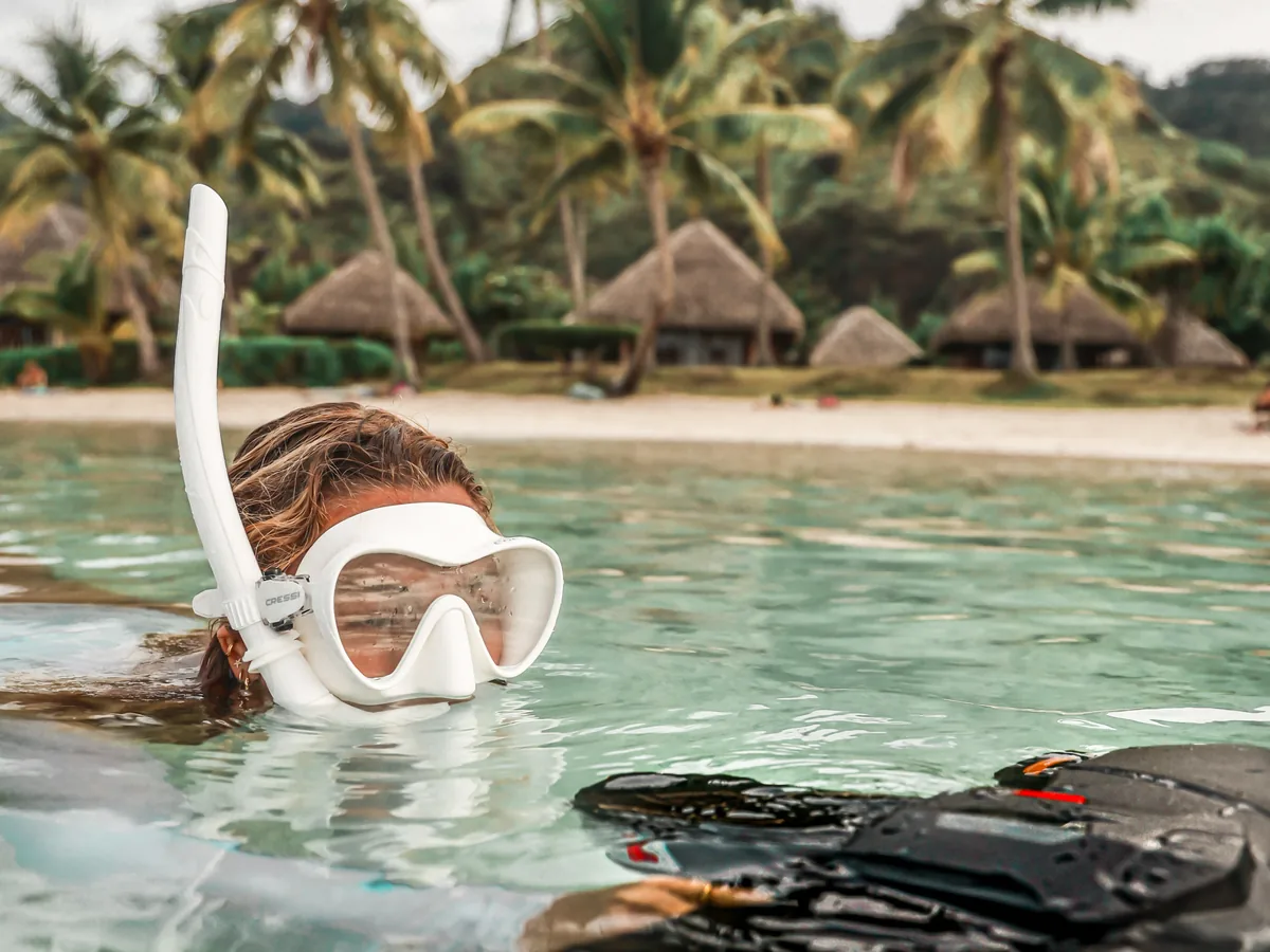 Cliente masquée dans l'eau peu profonde, son Sublue à côté d'elle, bungalows en arrière-plan