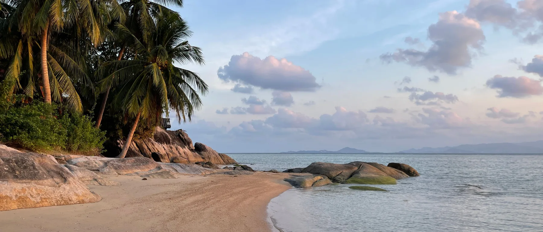 Plage de sable au petit matin, cocotiers penchés et rochers de granit